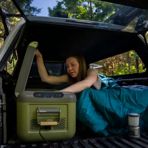 Woman using TRAVOCA portable car fridge inside a truck bed during outdoor camping, showing convenient cooling and compact design for overlanding adventures.