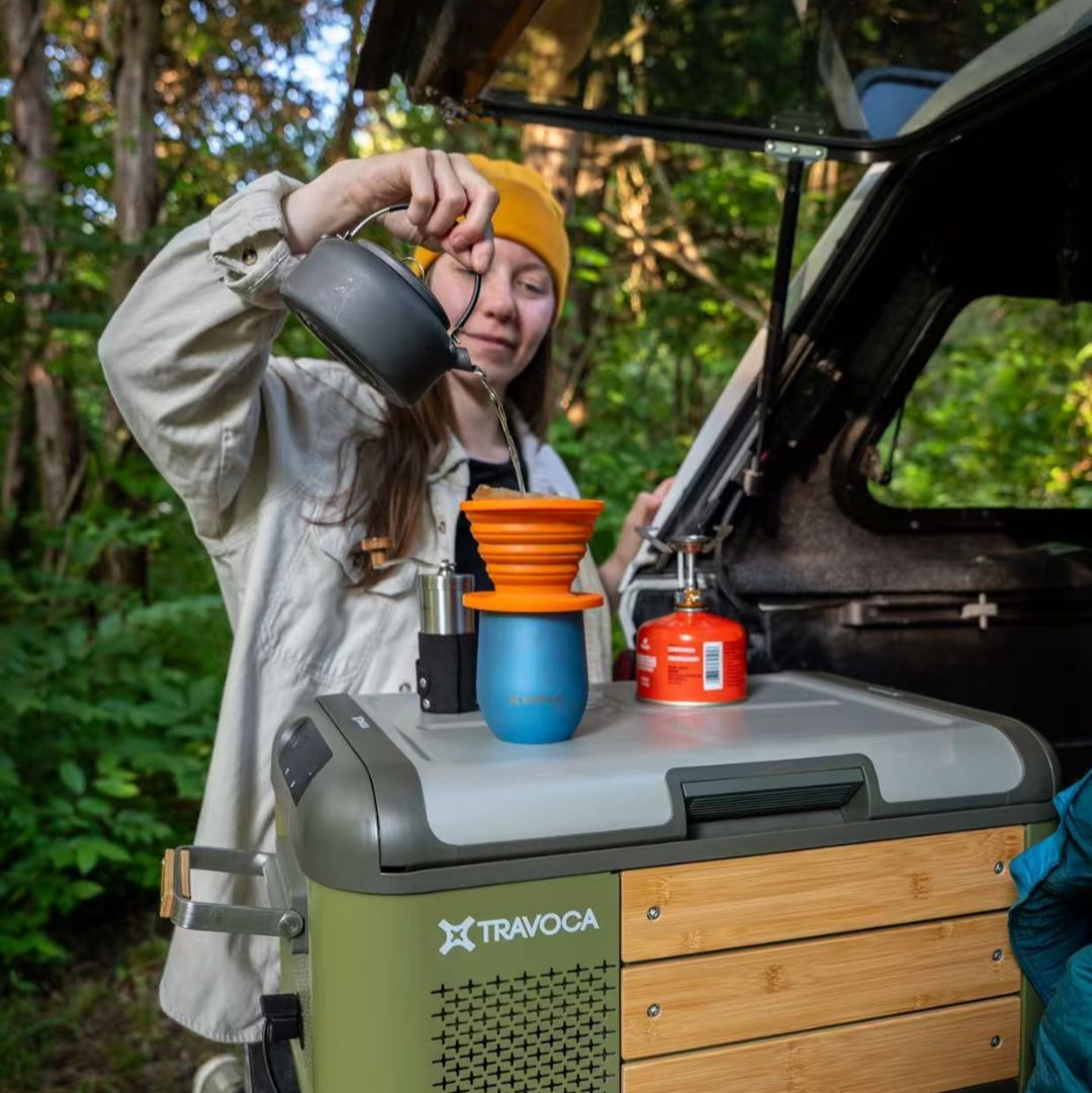 Female overlander making coffee beside TRAVOCA Rigel 45 dual zone ecooler during car camping trip, outdoor portable fridge for adventures.
